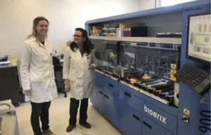 Two women in lab coats stand next to a large automated laboratory machine labeled "Bioerix" in a clinical laboratory setting.