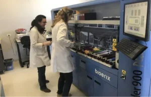 Two laboratory technicians in white coats work with automated equipment and blood sample vials in a clinical lab setting.