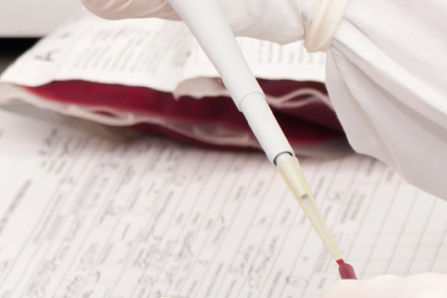A gloved hand uses a pipette to transfer liquid near paperwork and a blood bag on a laboratory table.