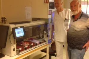 Two men stand beside a laboratory machine with test tubes and a computer screen displaying bar graphs in a clinical setting.
