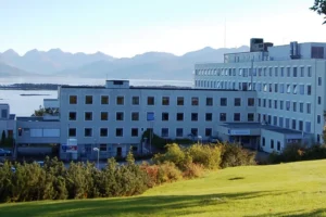 A multi-story hospital building with rectangular windows sits near a grassy area, with mountains and water visible in the background.