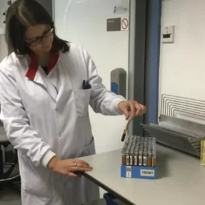 A person in a lab coat examines test tubes organized in a blue rack on a laboratory table.