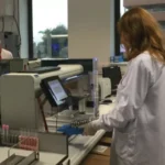 A person in a lab coat operates laboratory equipment, handling a tray of test tubes near a computer workstation.