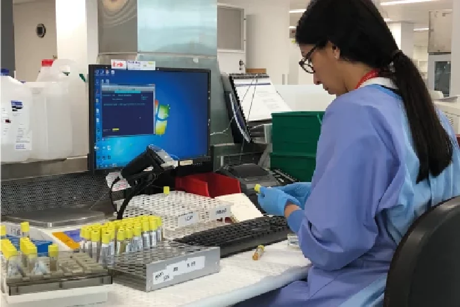 A lab technician wearing gloves and a lab coat handles test tubes at a workstation with a computer and various laboratory equipment.