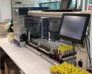 Automated laboratory equipment with a touchscreen interface processes multiple blood sample tubes, which are organized in racks on a workbench.