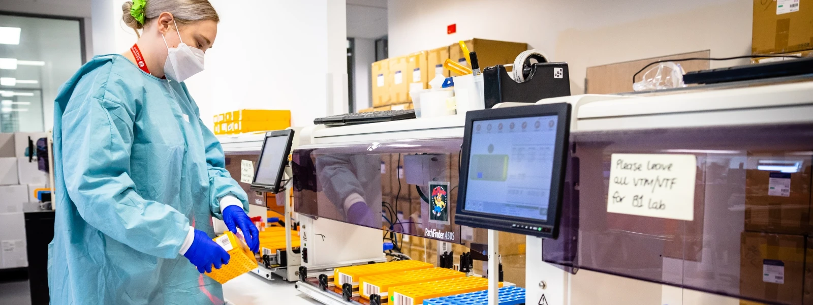A lab technician wearing protective gear handles orange sample racks beside automated laboratory equipment in a modern lab setting.