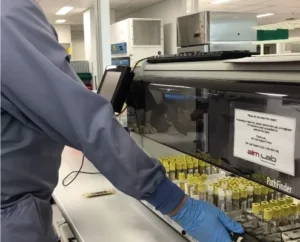 A person wearing gloves operates laboratory equipment, handling multiple test tubes with yellow caps in a clinical lab setting.