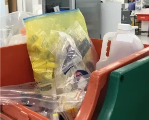 A plastic bin contains various items in clear plastic bags, including a gallon jug and yellow film canisters, at an airport security checkpoint.