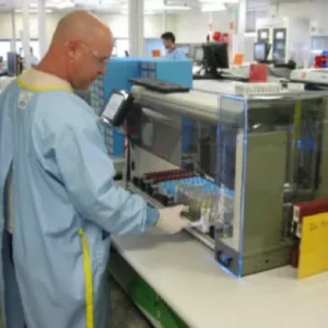 A lab technician in protective clothing operates an automated analyzer with multiple test tubes in a laboratory setting.