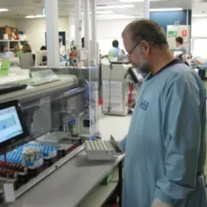 A laboratory technician in a blue lab coat operates equipment and handles samples in a busy laboratory setting.