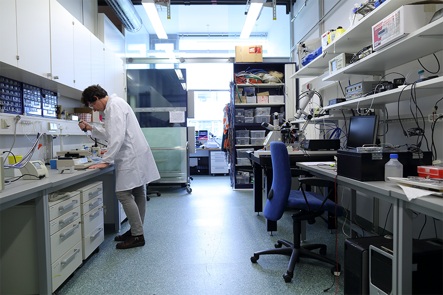 A person in a lab coat works with equipment at a laboratory workstation, conducting trace metals analysis; shelves and desks with scientific instruments and supplies are visible throughout the room.