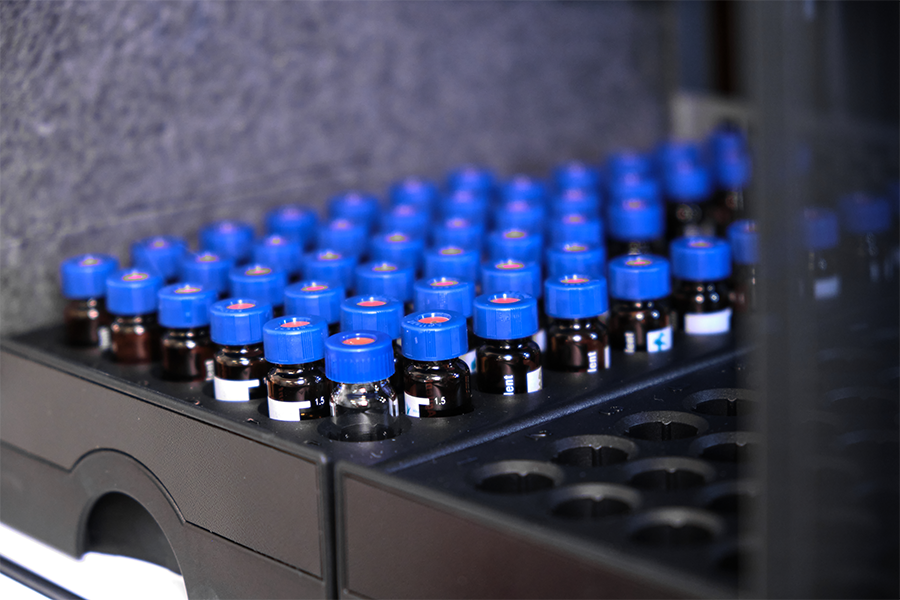 Rows of small glass vials with blue caps are arranged in a tray, likely in a laboratory setting, prepared for spectroscopy analysis.