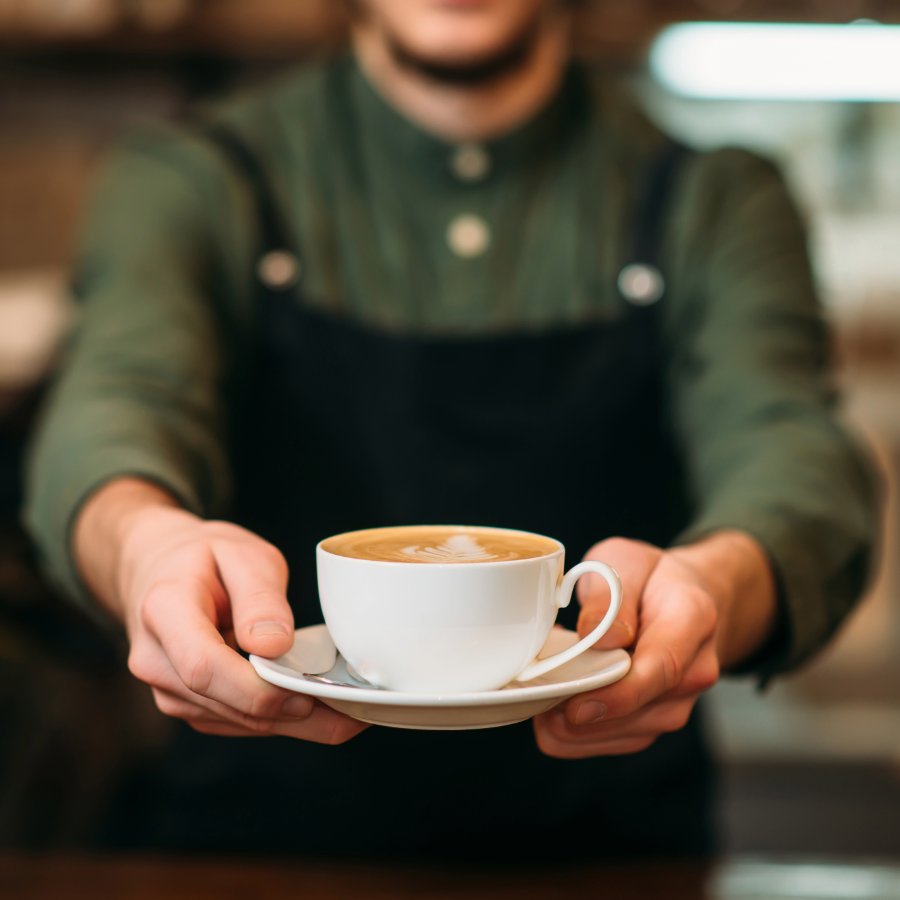 A barista in a green shirt and black apron presents a cup of cappuccino with latte art on a saucer, holding it out with both hands.